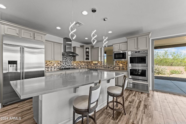 kitchen featuring hanging light fixtures, a kitchen island, wall chimney range hood, built in appliances, and hardwood / wood-style floors