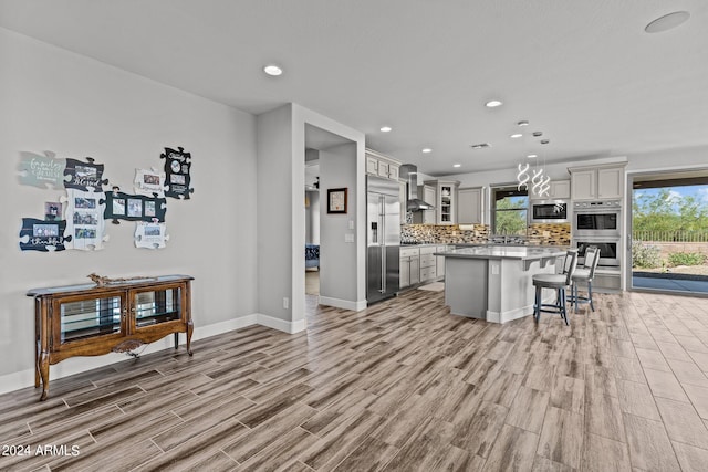 kitchen featuring built in appliances, wall chimney range hood, a kitchen island, light hardwood / wood-style flooring, and decorative light fixtures