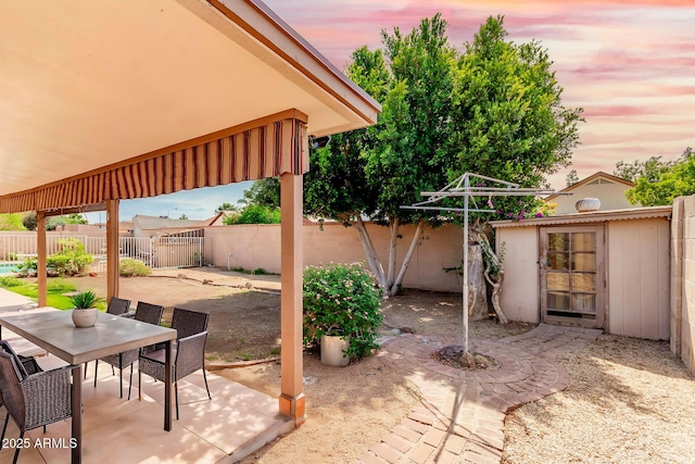 patio terrace at dusk with a fenced backyard, outdoor dining area, and an outdoor structure