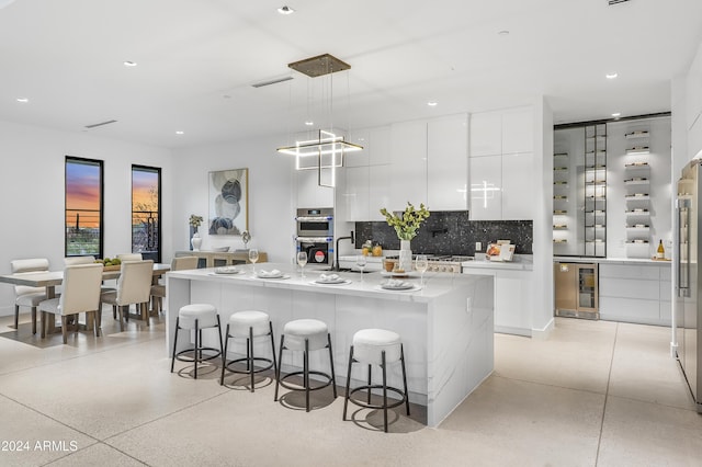 kitchen featuring white cabinetry, hanging light fixtures, tasteful backsplash, wine cooler, and an island with sink