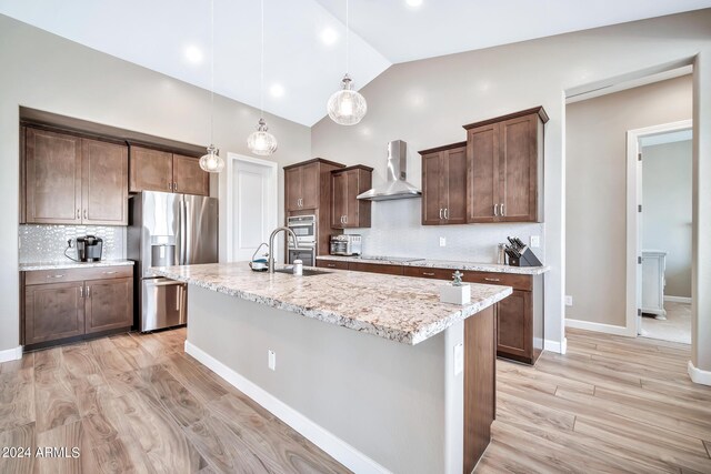 kitchen featuring a kitchen island with sink, wall chimney range hood, sink, hanging light fixtures, and double oven