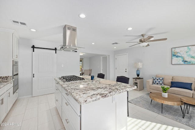 kitchen featuring ceiling fan, island exhaust hood, light stone counters, a barn door, and white cabinetry