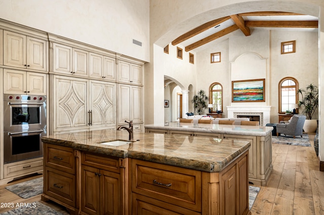 kitchen with an island with sink, light wood-type flooring, beamed ceiling, stone counters, and double oven