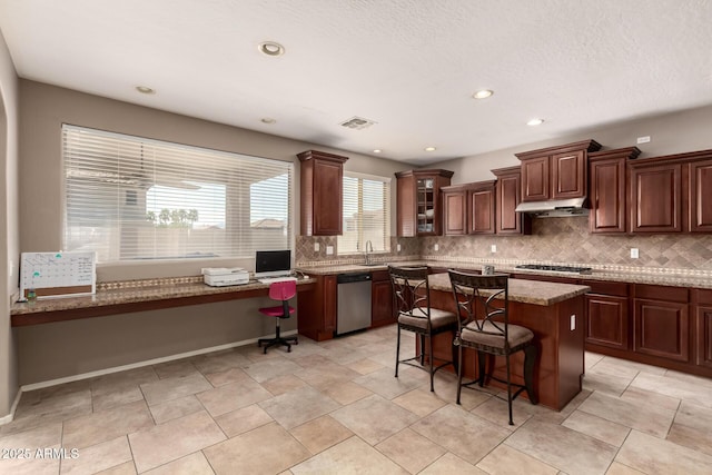 kitchen featuring built in study area, stainless steel appliances, under cabinet range hood, a kitchen bar, and backsplash