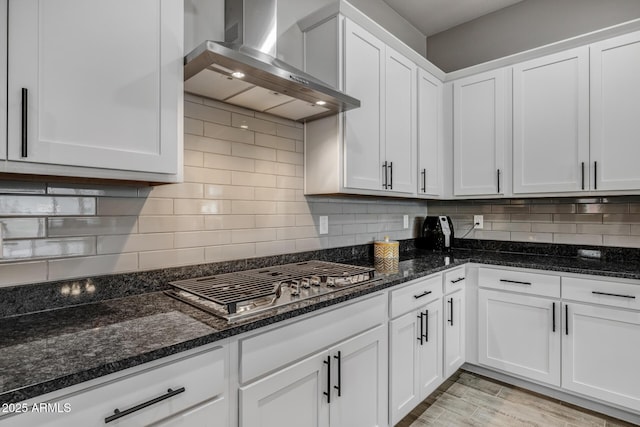 kitchen with wall chimney range hood, stainless steel gas stovetop, dark stone counters, and white cabinets