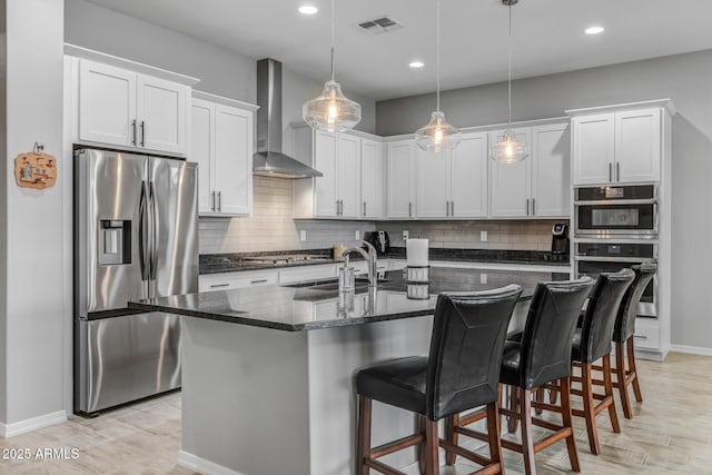 kitchen with stainless steel appliances, a center island with sink, wall chimney range hood, and white cabinets