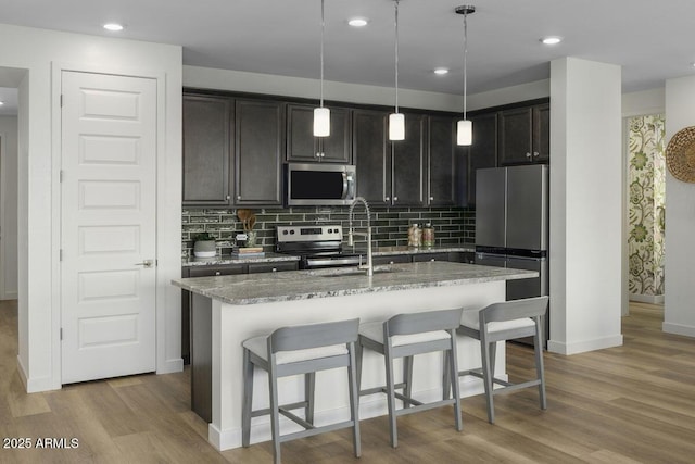 kitchen featuring pendant lighting, light hardwood / wood-style flooring, stainless steel appliances, light stone countertops, and an island with sink