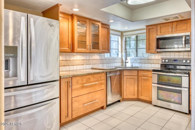 kitchen featuring appliances with stainless steel finishes, a sink, tasteful backsplash, glass insert cabinets, and light stone counters