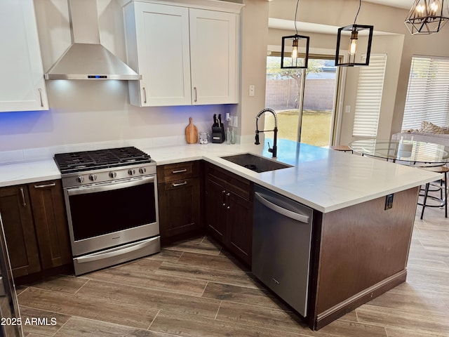 kitchen with white cabinets, appliances with stainless steel finishes, a peninsula, wall chimney range hood, and a sink