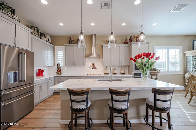 kitchen featuring hanging light fixtures, an island with sink, stainless steel appliances, and wall chimney range hood