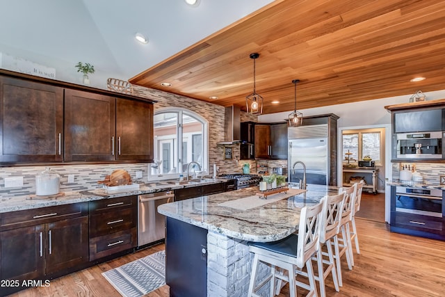 kitchen featuring stainless steel appliances, tasteful backsplash, an island with sink, light stone countertops, and wood ceiling