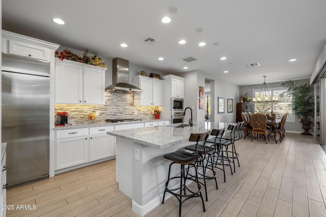 kitchen featuring white cabinets, built in appliances, a kitchen island with sink, and wall chimney range hood