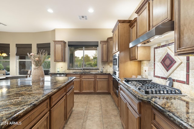 2378 N 158th Dr, Goodyear, AZ 85395 - kitchen featuring appliances with stainless steel finishes, decorative backsplash, brown cabinetry, dark stone counters, and under cabinet range hood