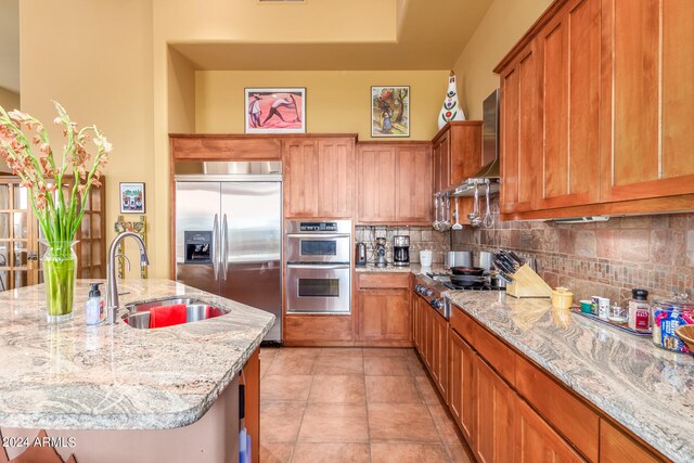 kitchen with stainless steel appliances, tasteful backsplash, brown cabinetry, a sink, and wall chimney range hood