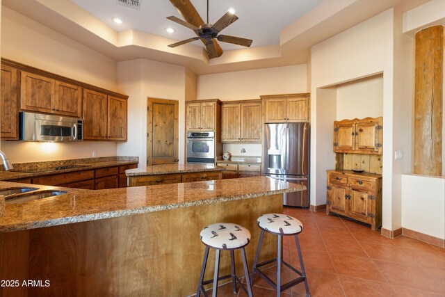 kitchen featuring a raised ceiling, dark tile patterned flooring, brown cabinets, stainless steel appliances, and stone counters