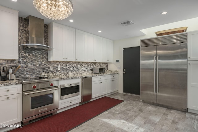 kitchen with wall chimney range hood, white cabinets, light stone counters, and high end appliances