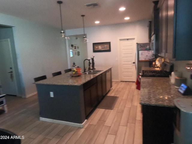 kitchen with decorative light fixtures, an island with sink, sink, light stone counters, and light wood-type flooring