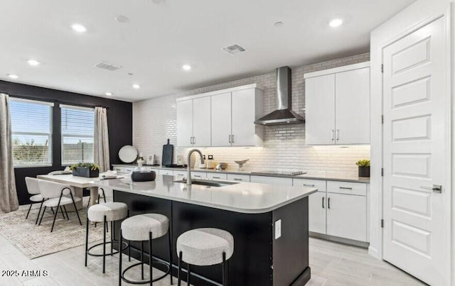 kitchen featuring an island with sink, light countertops, wall chimney range hood, white cabinetry, and a sink