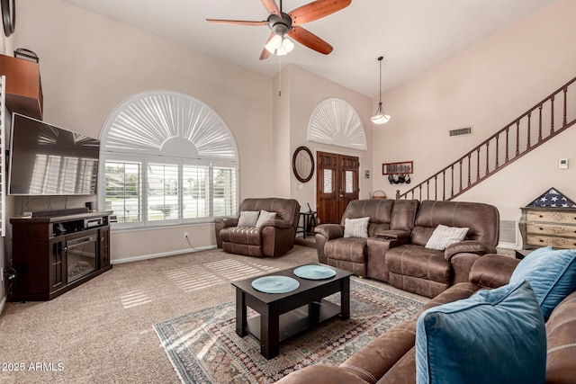 carpeted living room with stairway, a ceiling fan, baseboards, visible vents, and a towering ceiling