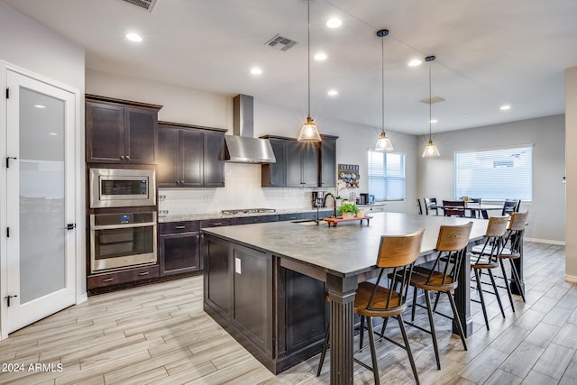 kitchen featuring pendant lighting, dark brown cabinets, a center island with sink, wall chimney range hood, and appliances with stainless steel finishes