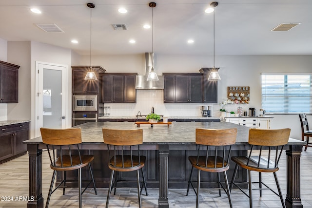 kitchen with decorative light fixtures, wall chimney exhaust hood, appliances with stainless steel finishes, a kitchen breakfast bar, and a spacious island