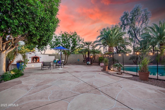 patio terrace at dusk featuring a fenced in pool, a fenced backyard, and a lit fireplace