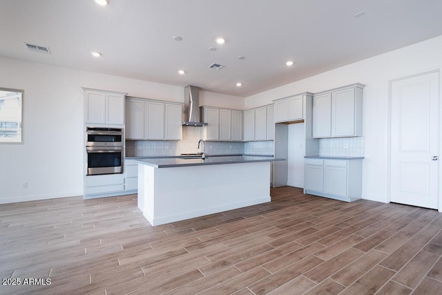 876 W Golden Ore Pass, Apache Junction, AZ 85120 - kitchen featuring wall chimney exhaust hood, double oven, light wood finished floors, backsplash, and a kitchen island with sink