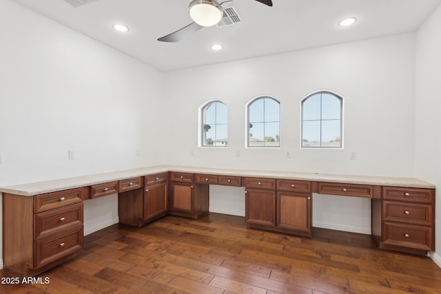 unfurnished office featuring plenty of natural light, dark hardwood / wood-style floors, built in desk, and ceiling fan