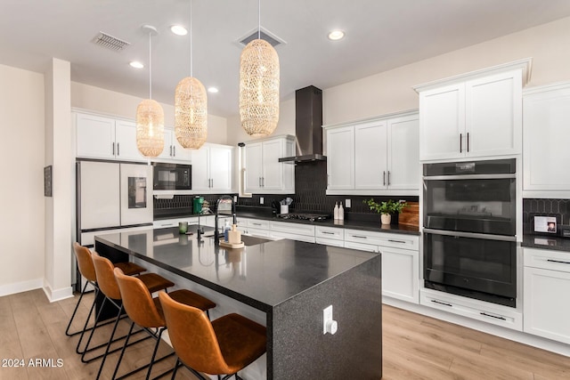 kitchen with pendant lighting, black appliances, a center island with sink, wall chimney range hood, and light wood-type flooring