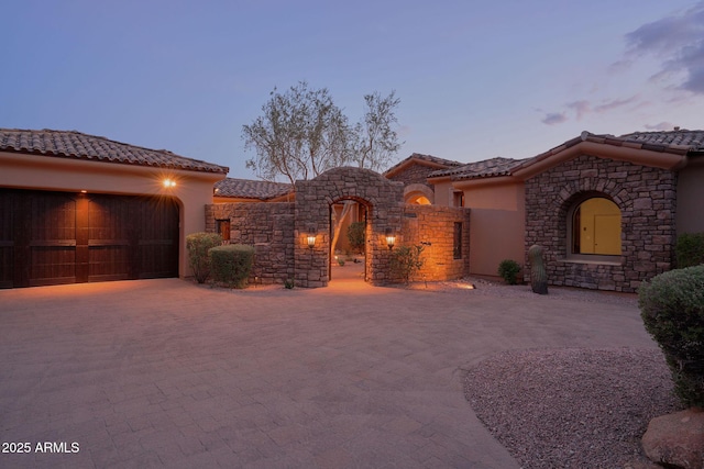 mediterranean / spanish-style home with stone siding, a garage, a tiled roof, stucco siding, and decorative driveway