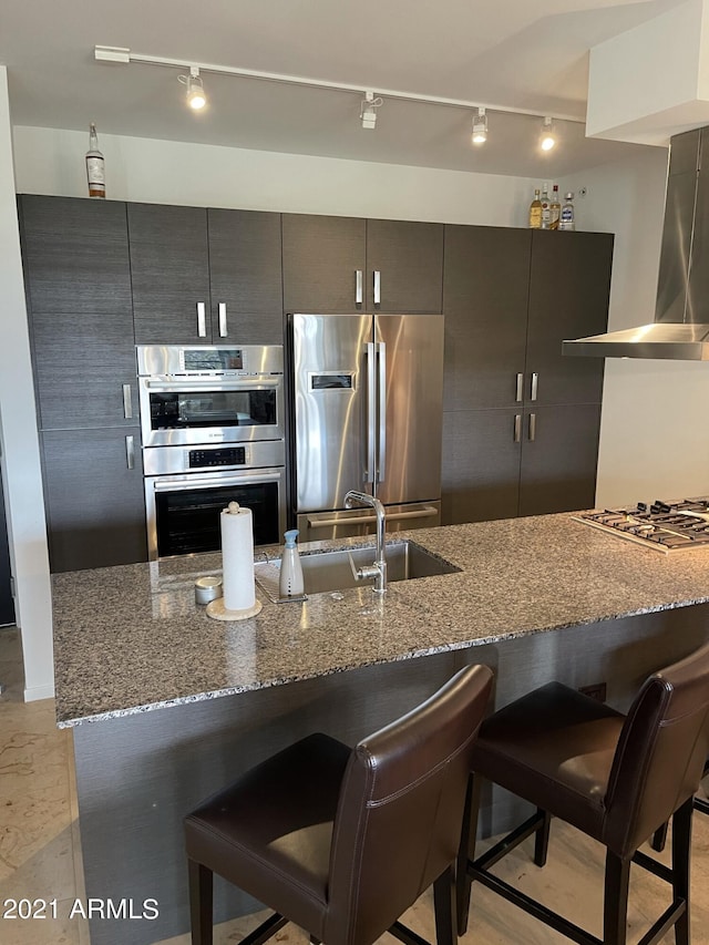 kitchen with dark stone countertops, a sink, a kitchen breakfast bar, stainless steel appliances, and wall chimney exhaust hood