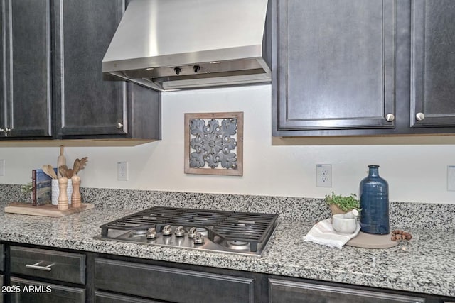 kitchen with wall chimney range hood, dark cabinets, light stone counters, and stainless steel gas stovetop