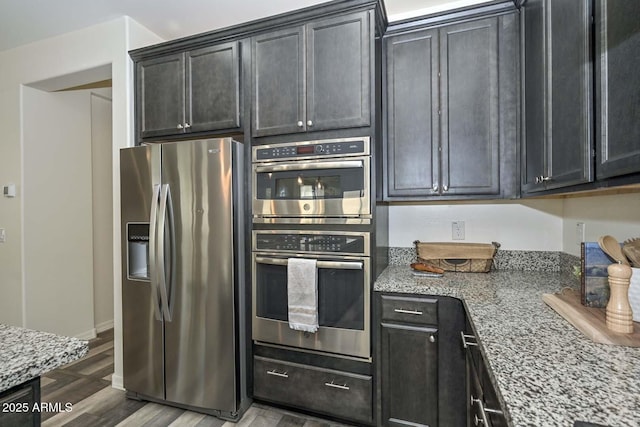 kitchen featuring stainless steel appliances, dark cabinetry, light stone counters, and wood finished floors