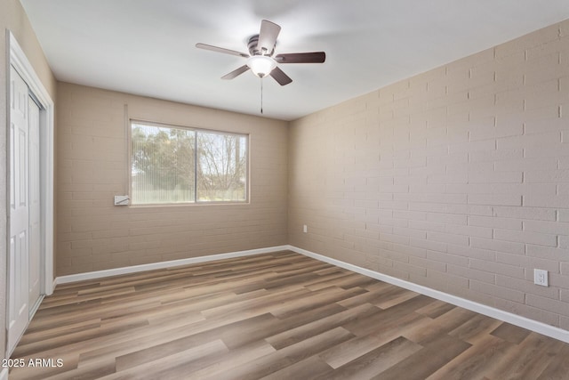 empty room featuring a ceiling fan, brick wall, baseboards, and wood finished floors