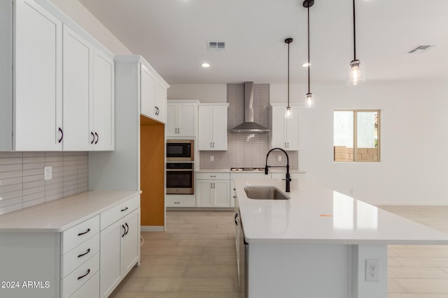 kitchen featuring pendant lighting, stainless steel oven, a kitchen island with sink, wall chimney range hood, and sink