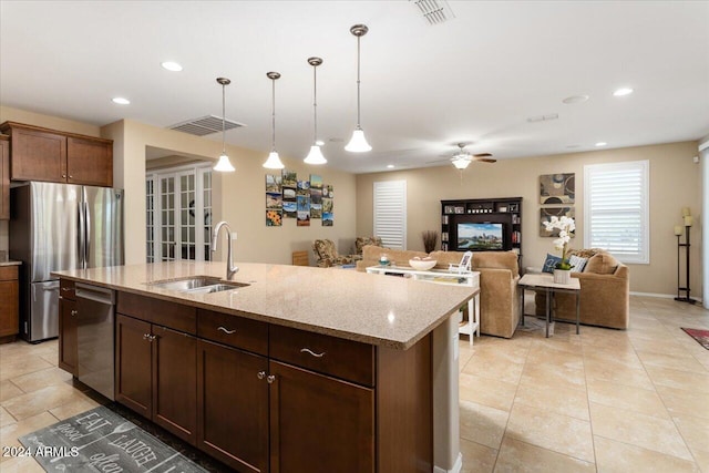 kitchen with stainless steel appliances, a center island with sink, sink, light stone countertops, and decorative light fixtures