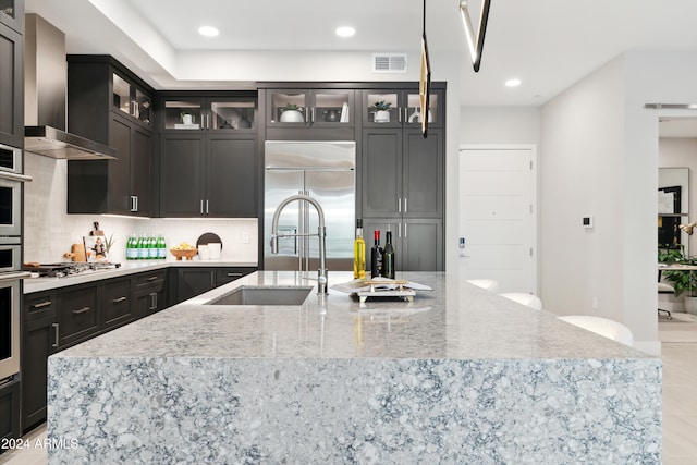 kitchen featuring sink, wall chimney range hood, light stone counters, an island with sink, and appliances with stainless steel finishes