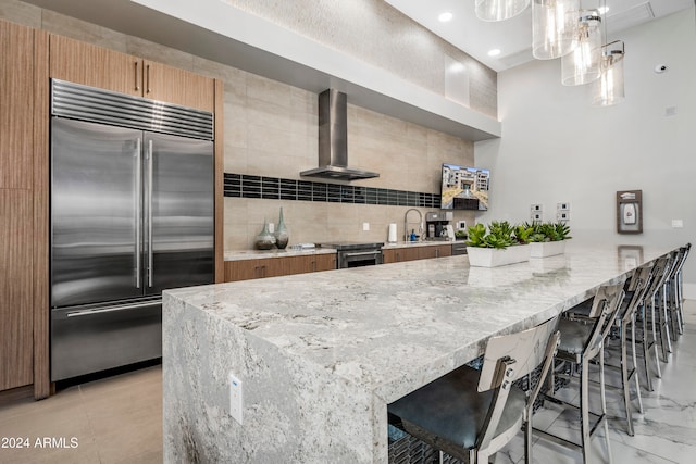 kitchen with wall chimney exhaust hood, stainless steel appliances, backsplash, decorative light fixtures, and a kitchen bar