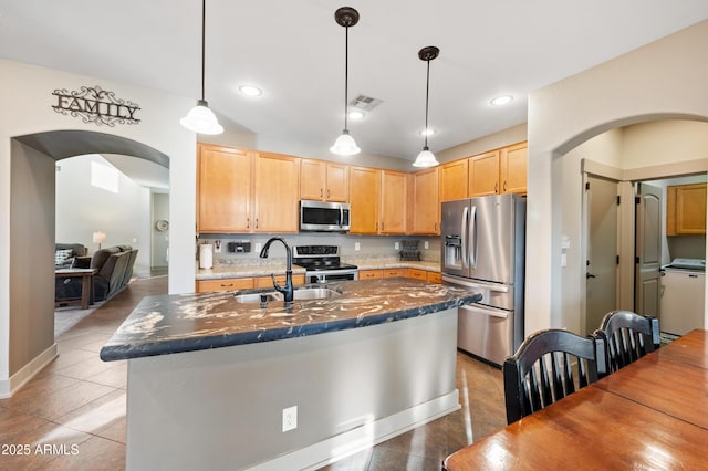 kitchen featuring light brown cabinetry, washer / dryer, appliances with stainless steel finishes, pendant lighting, and a kitchen island with sink