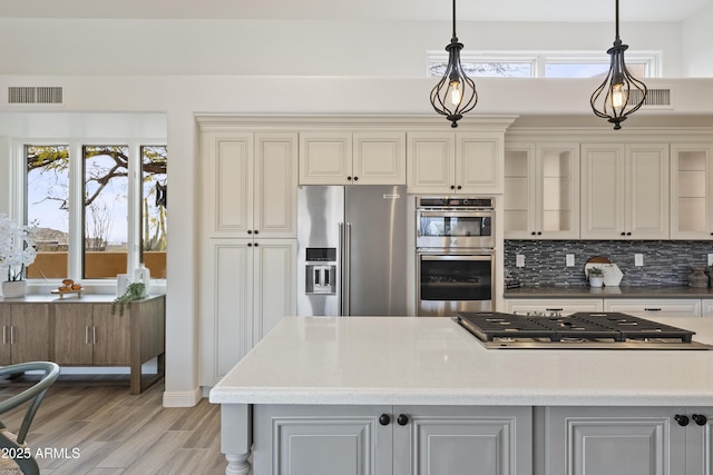 kitchen featuring pendant lighting, stainless steel appliances, visible vents, backsplash, and glass insert cabinets