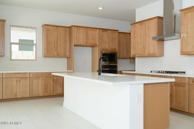 kitchen featuring an island with sink, tasteful backsplash, gas stovetop, wall chimney exhaust hood, and light countertops