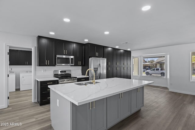 kitchen with washer and dryer, a center island with sink, sink, light stone countertops, and appliances with stainless steel finishes