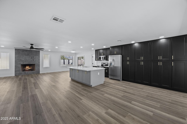 kitchen featuring a center island with sink, appliances with stainless steel finishes, light wood-type flooring, a tile fireplace, and sink