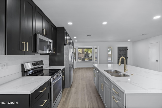 kitchen featuring an island with sink, appliances with stainless steel finishes, light stone countertops, light hardwood / wood-style flooring, and sink