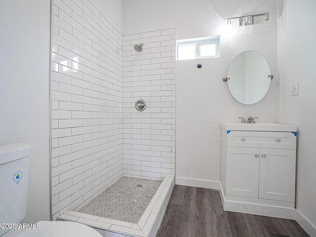 bathroom with toilet, wood finished floors, vanity, baseboards, and a tile shower