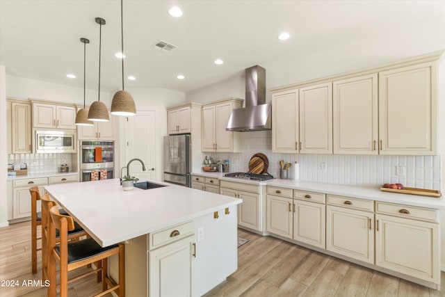 kitchen with sink, cream cabinets, a center island with sink, wall chimney exhaust hood, and stainless steel appliances