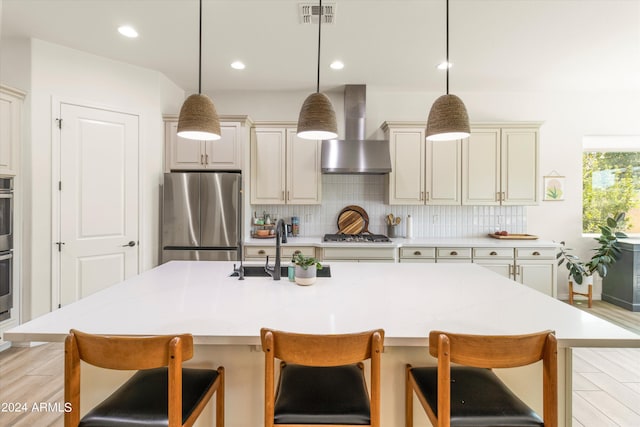 kitchen with cream cabinets, a center island with sink, wall chimney exhaust hood, appliances with stainless steel finishes, and a kitchen breakfast bar