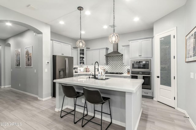 kitchen with a sink, stainless steel appliances, arched walkways, wall chimney exhaust hood, and light countertops