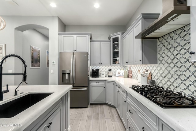 kitchen with stainless steel refrigerator with ice dispenser, black gas stovetop, a sink, wall chimney exhaust hood, and decorative backsplash