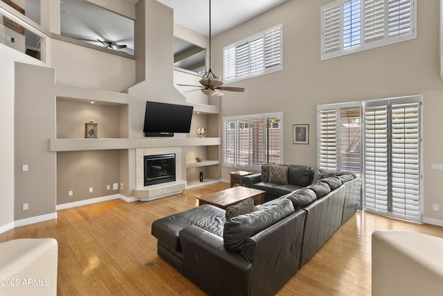 living room with ceiling fan, a tiled fireplace, baseboards, wood finished floors, and a towering ceiling